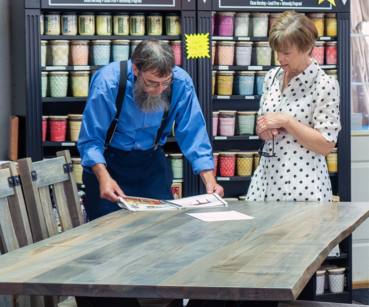 Stacks of raw hardwood planks on a cart in an Amish woodworking shop, with more wood stacks visible in the background.