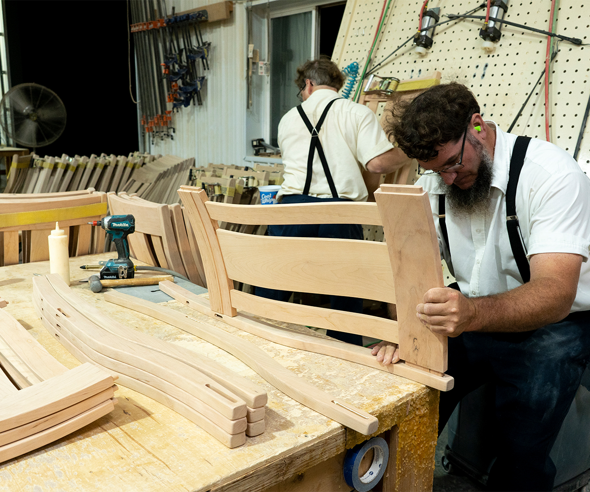 Craftsman assembling custom furniture chair back in busy woodshop with tools and curved hardwood pieces.