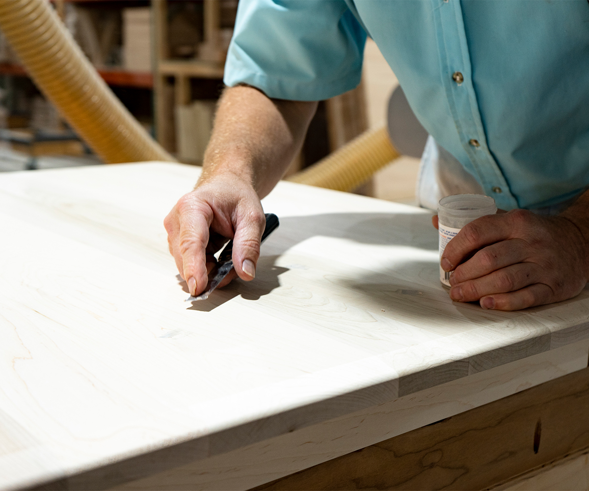Craftsman detailing custom furniture tabletop, filling small imperfections on light hardwood surface.