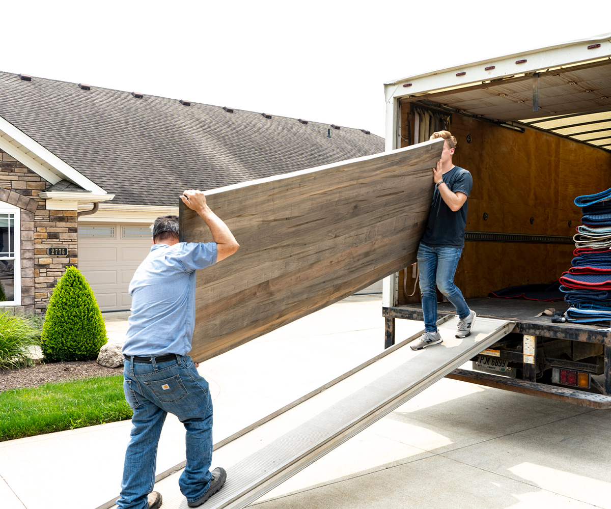 Amish furniture delivery team carrying large tabletop from truck into suburban home.